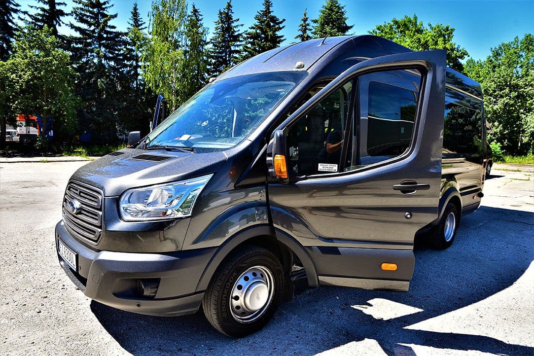 Szary Ford Transit z otwartymi drzwiami bocznymi, stojący na parkingu wśród drzew.
